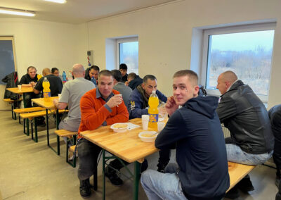 Skupina ľudí sediacich za dlhými stolmi v jedálni pri spoločnom obede.